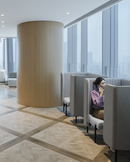 Modern office space with large windows, sleek wooden flooring, and a woman sitting in a private lounge booth talking on a phone.
