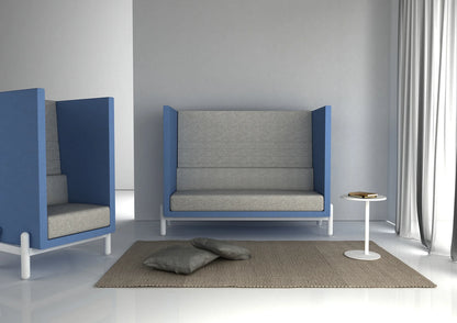 Modern office seating area with blue privacy pods, gray cushions, a beige rug, and a round white side table holding books.